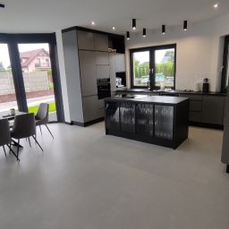 GW Projekt Grzegorz Worobiej - Nowoczesna kuchnia z wyspą i posadzką z mikrocementu w szarościach. Minimalistyczny design, duże okna, oświetlenie punktowe. Widok na zewnątrz.