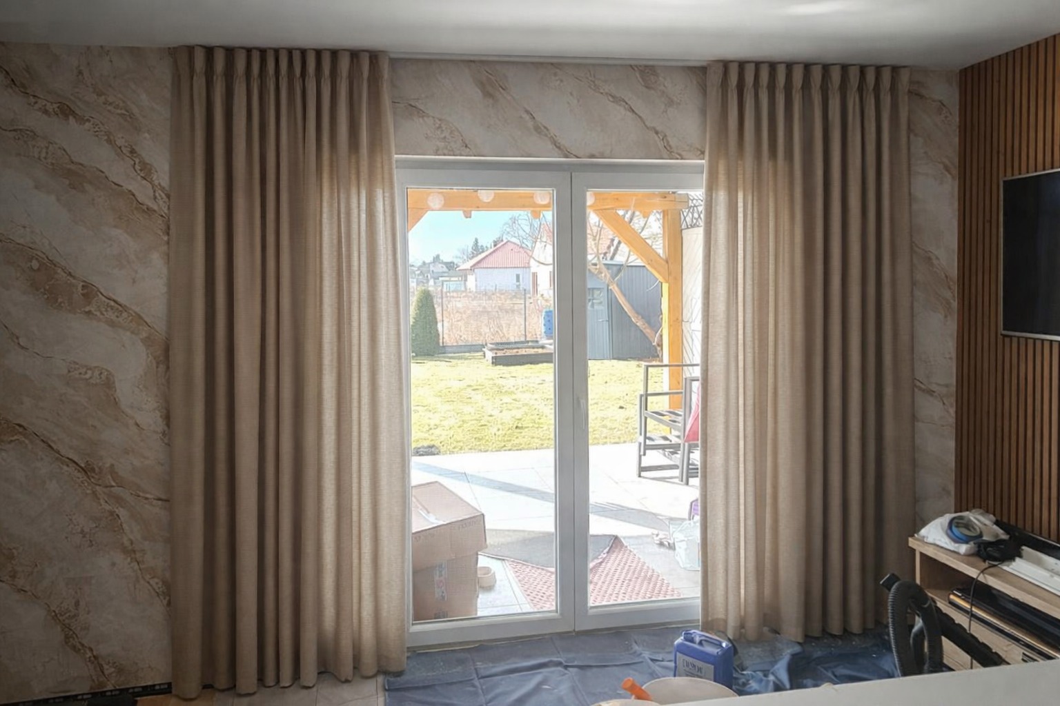Beżowe, gładkie zasłony na szynie, z delikatnym marszczeniem, wiszące po obu stronach białych drzwi balkonowych. W tle widoczny ogród i fragment elewacji domu.