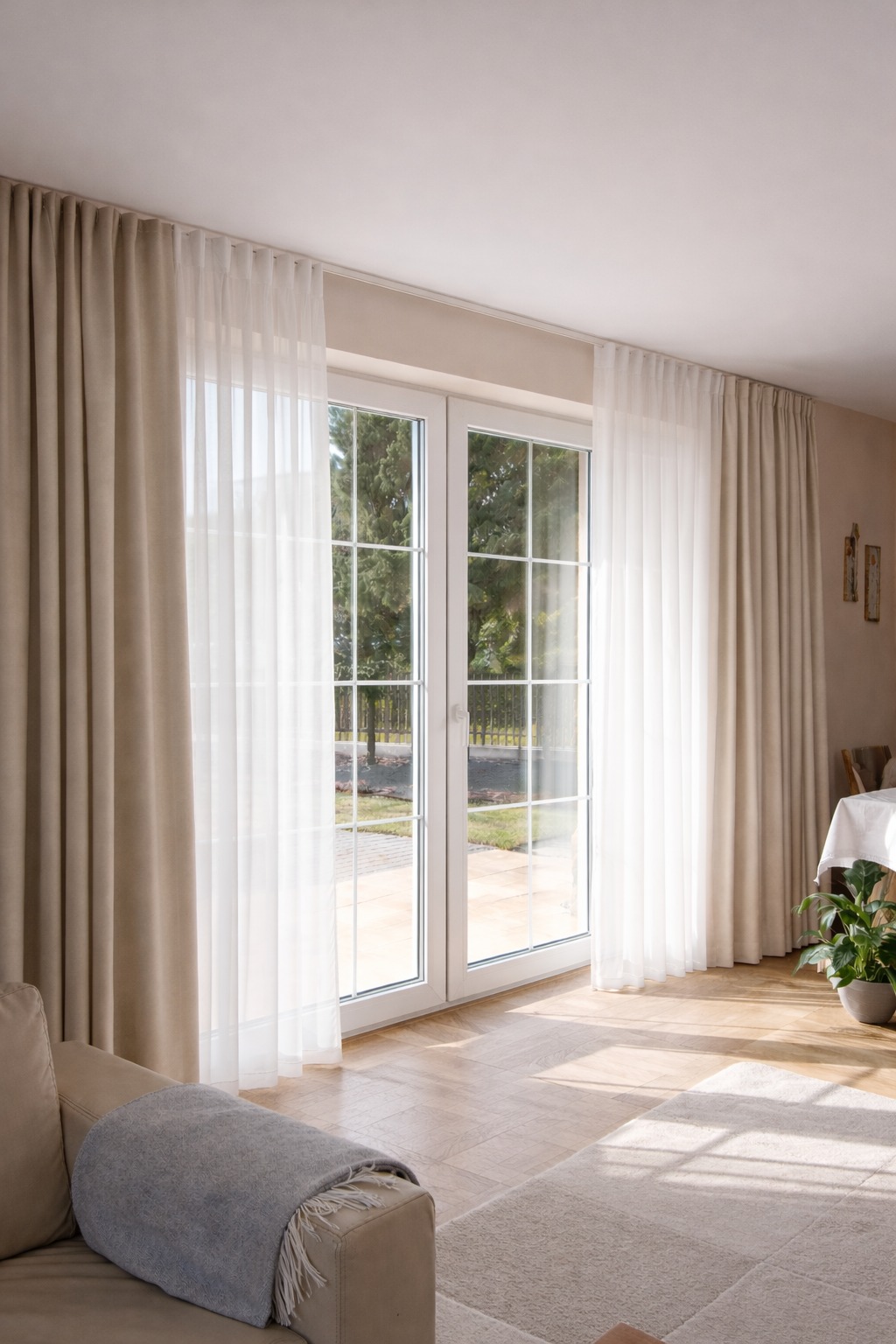 Jasne, beżowe zasłony i firany w salonie z dużym oknem balkonowym. Naturalne światło, parkiet i dywan tworzą przytulną atmosferę. Widok na zieleń za oknem.