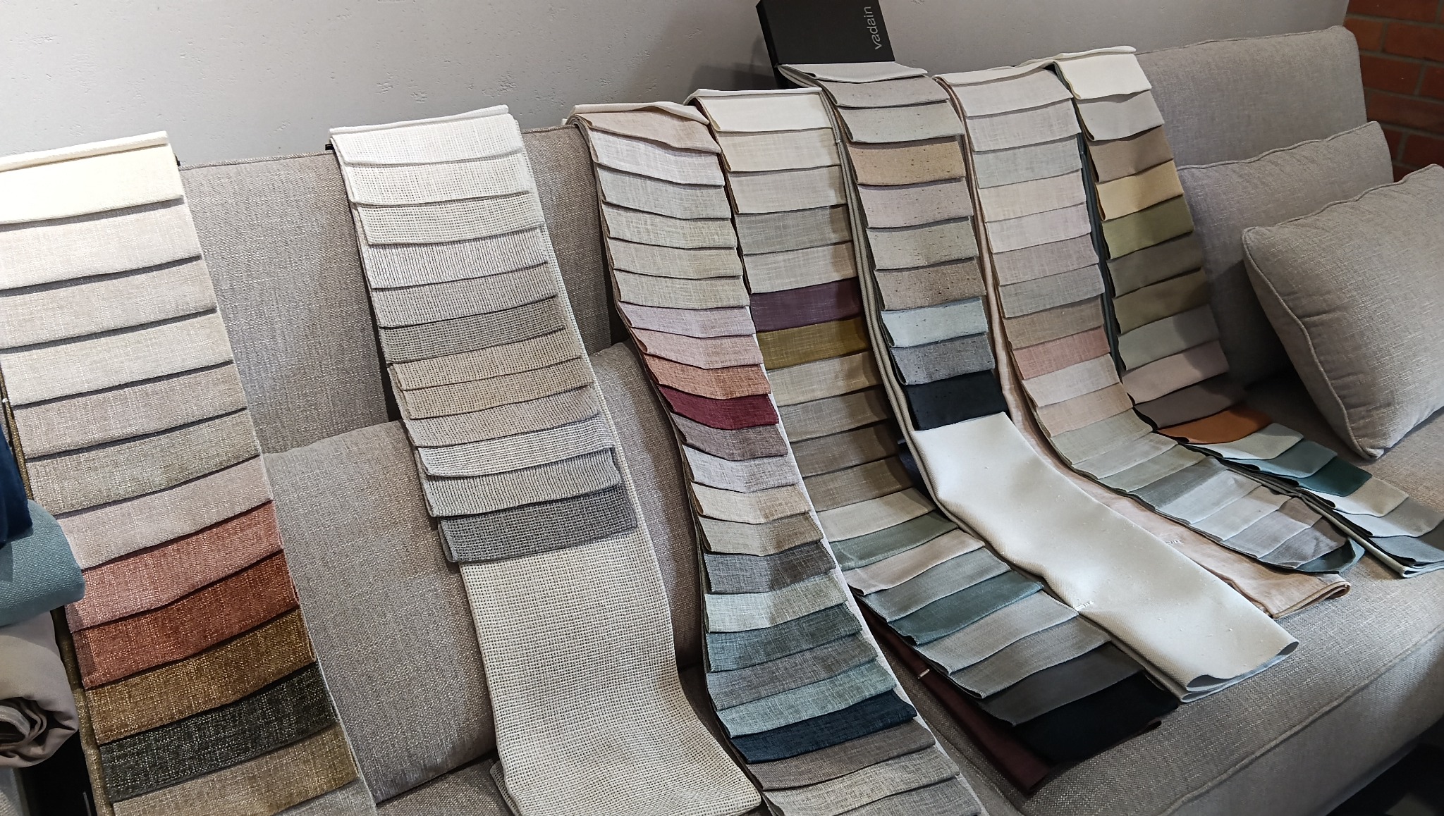 Próbniki tkanin w różnych odcieniach szarości, beżu, brązu i zieleni, ułożone na szarej sofie z poduszkami. Widoczne faktury materiałów i subtelne różnice kolorystyczne.