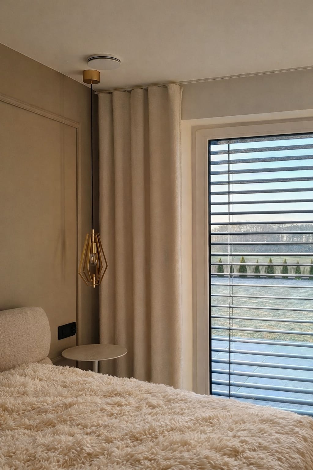 Beżowa zasłona w eleganckim wnętrzu z widokiem na zewnątrz przez żaluzje. Złota lampa wisząca dodaje szyku. Przytulne łóżko z miękką narzutą.