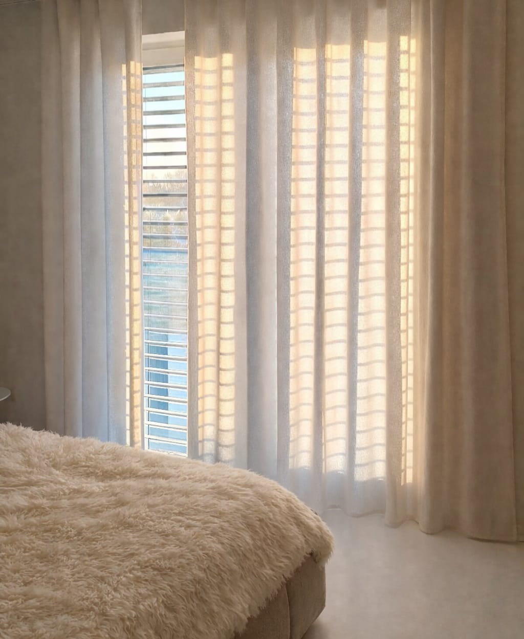 Delikatne, beżowe zasłony z lnu, przepuszczające światło, tworzą subtelny wzór cienia na tle okna z żaluzjami. Widoczny fragment łóżka z puszystą narzutą.