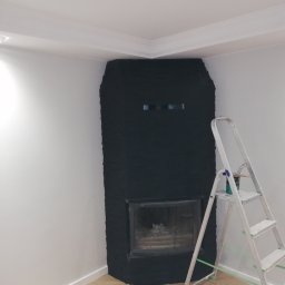 Jacek Szczepański - Świeżo pomalowany na czarno kominek z cegły w rogu pokoju, drabina malarska z farbą obok. Widoczne zabezpieczenia podłogi. Białe ściany i sufit z dekoracyjną listwą.