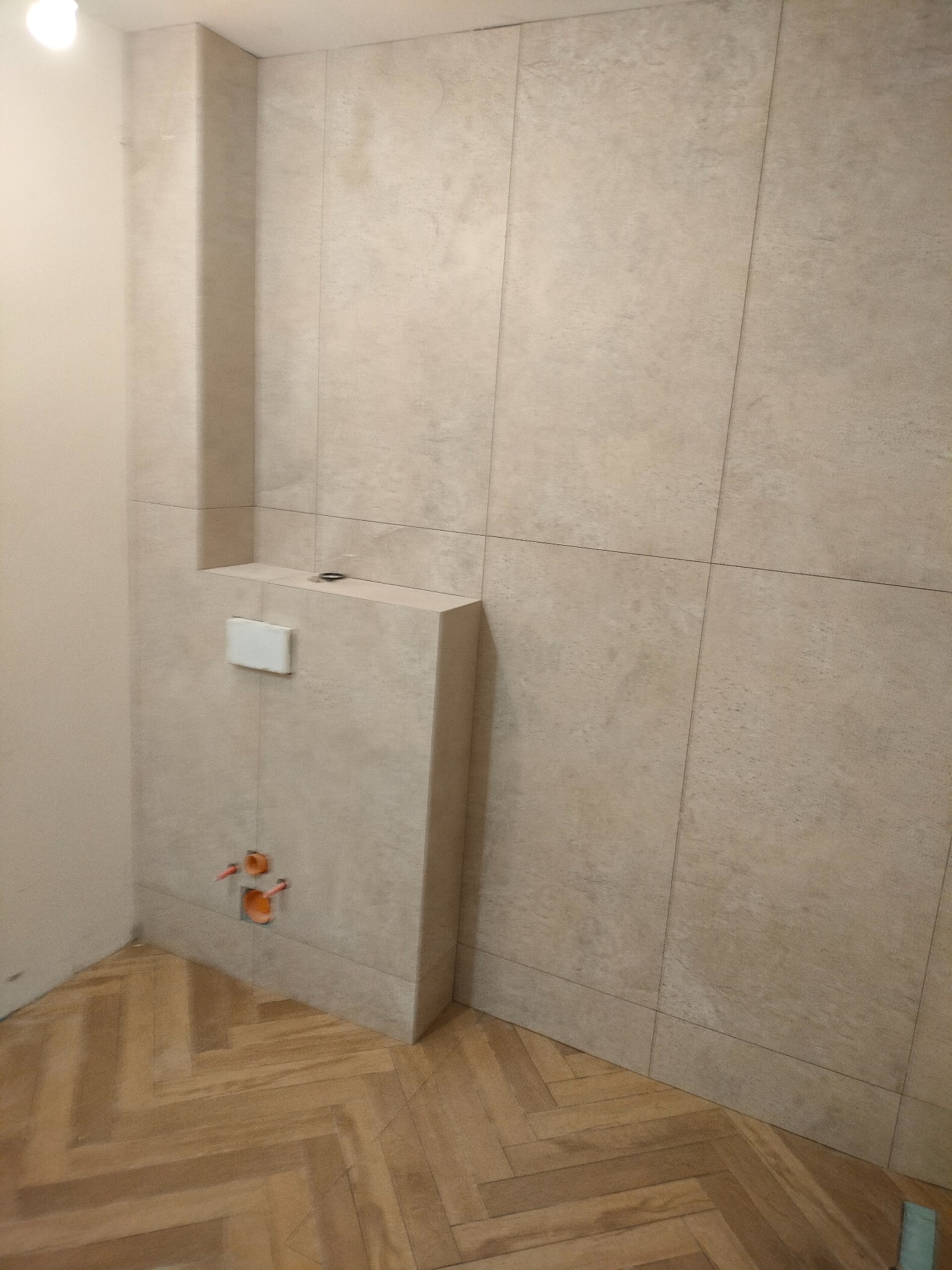 Wykończona łazienka z beżowymi płytkami imitującymi kamień, drewnianą podłogą w jodełkę i zabudową podtynkową WC z białym przyciskiem. Widoczne przyłącza wodne.