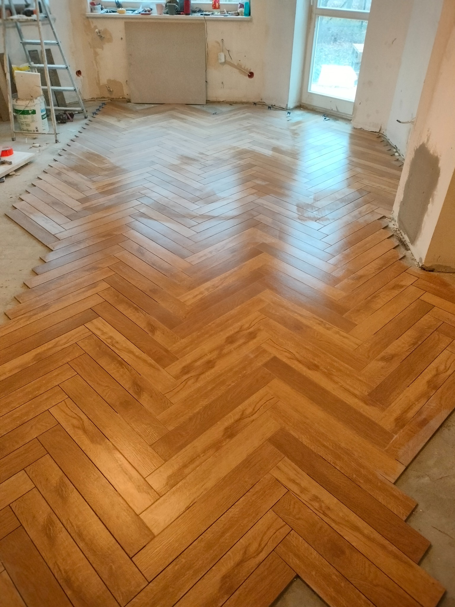 Układanie parkietu w jodełkę w jasnym wnętrzu. Widoczne niedokończone krawędzie i drabina malarska w tle. Parkiet z wyraźnym rysunkiem drewna.