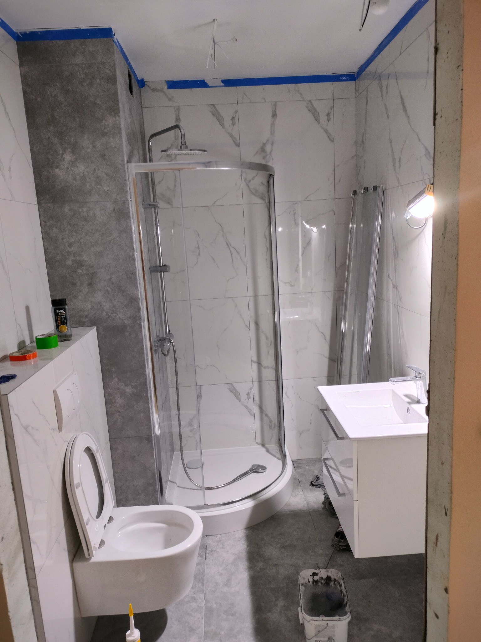 Wnętrze łazienki w trakcie wykańczania: kabina prysznicowa, umywalka z szafką, podwieszana toaleta. Widoczne materiały i narzędzia glazurnicze.