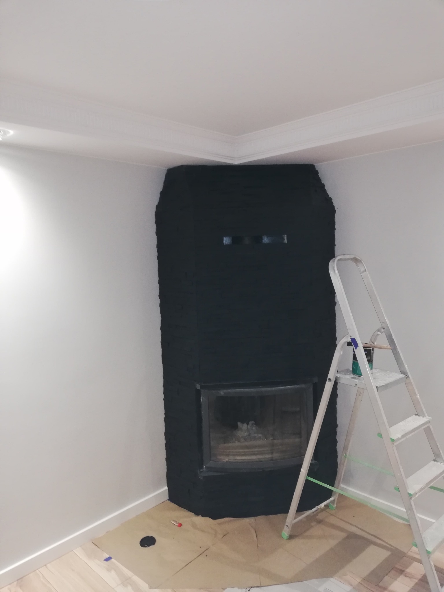 Świeżo pomalowany na czarno kominek z cegły w rogu pokoju, drabina malarska z farbą obok. Widoczne zabezpieczenia podłogi. Białe ściany i sufit z dekoracyjną listwą.