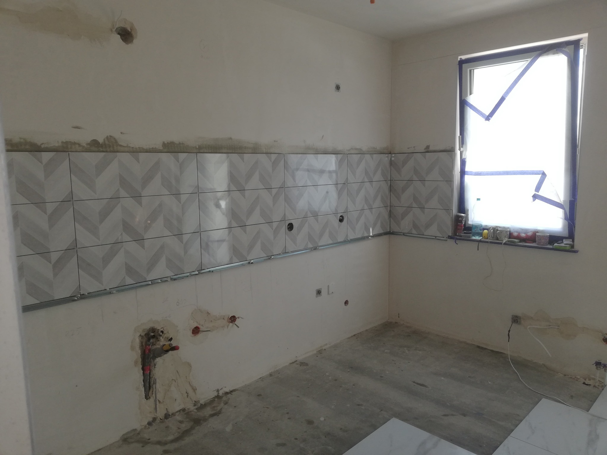 Montaż płytek w kuchni: wzór w jodełkę na ścianie, widoczne niedokończone prace, surowy stan podłogi, okno zabezpieczone folią malarską.