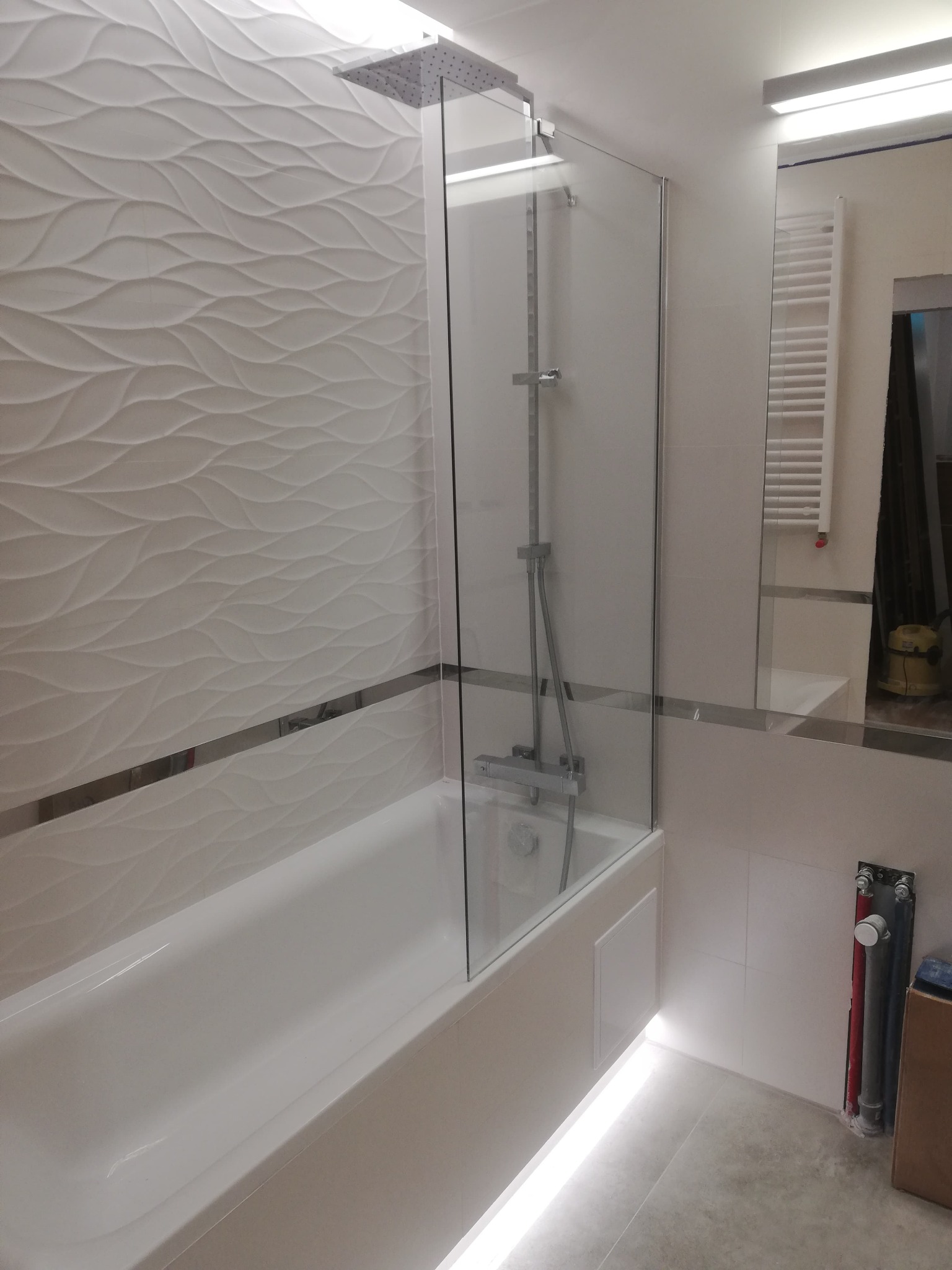 Nowoczesna łazienka po remoncie: biała wanna z parawanem, dekoracyjne płytki 3D na ścianie, oświetlenie LED pod wanną, grzejnik drabinkowy w lustrze.