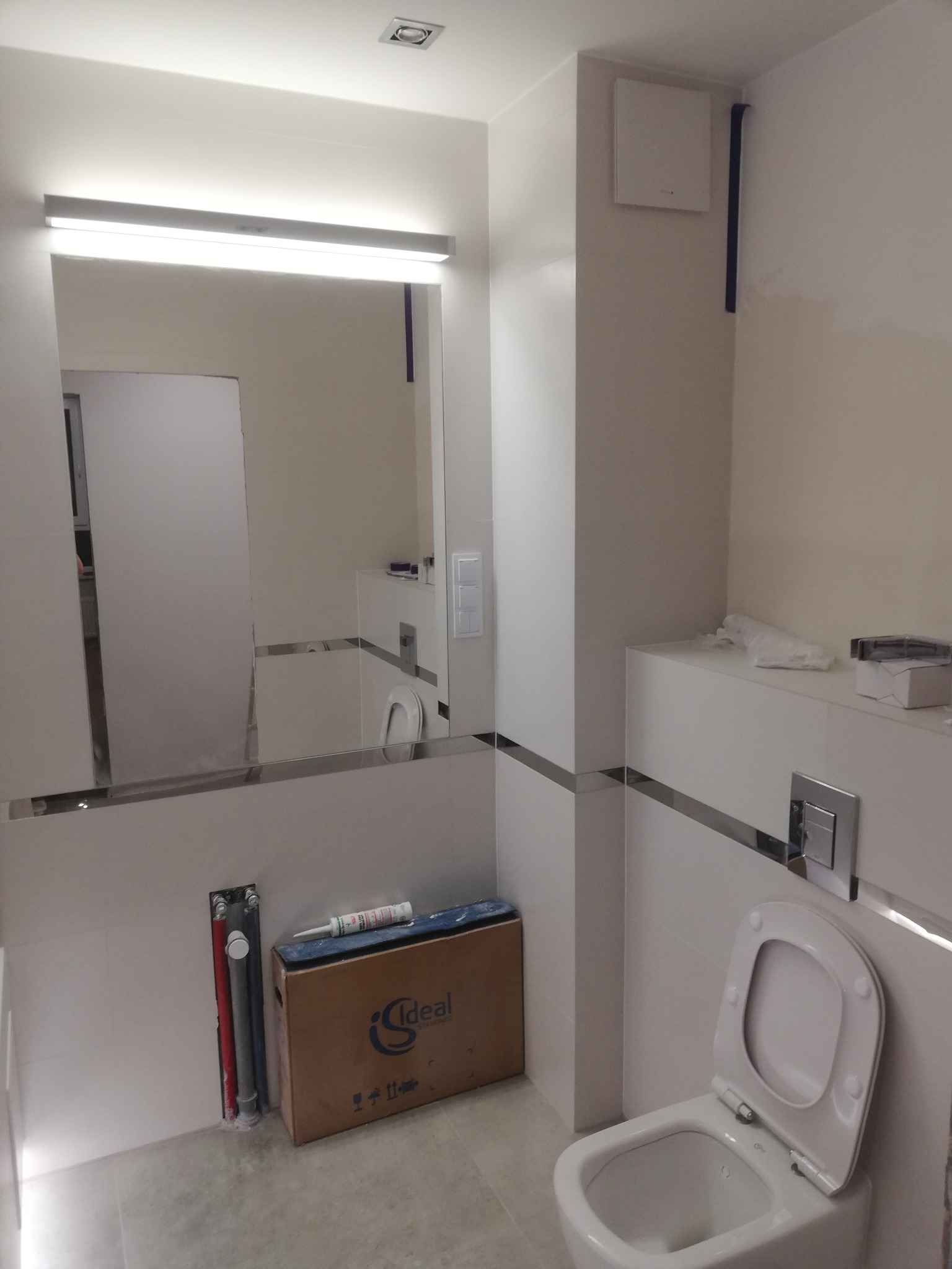 Nowoczesna łazienka w bieli z toaletą, lustrem z podświetleniem i elementami wykończenia w chromie. Widoczne narzędzia i materiały instalacyjne.