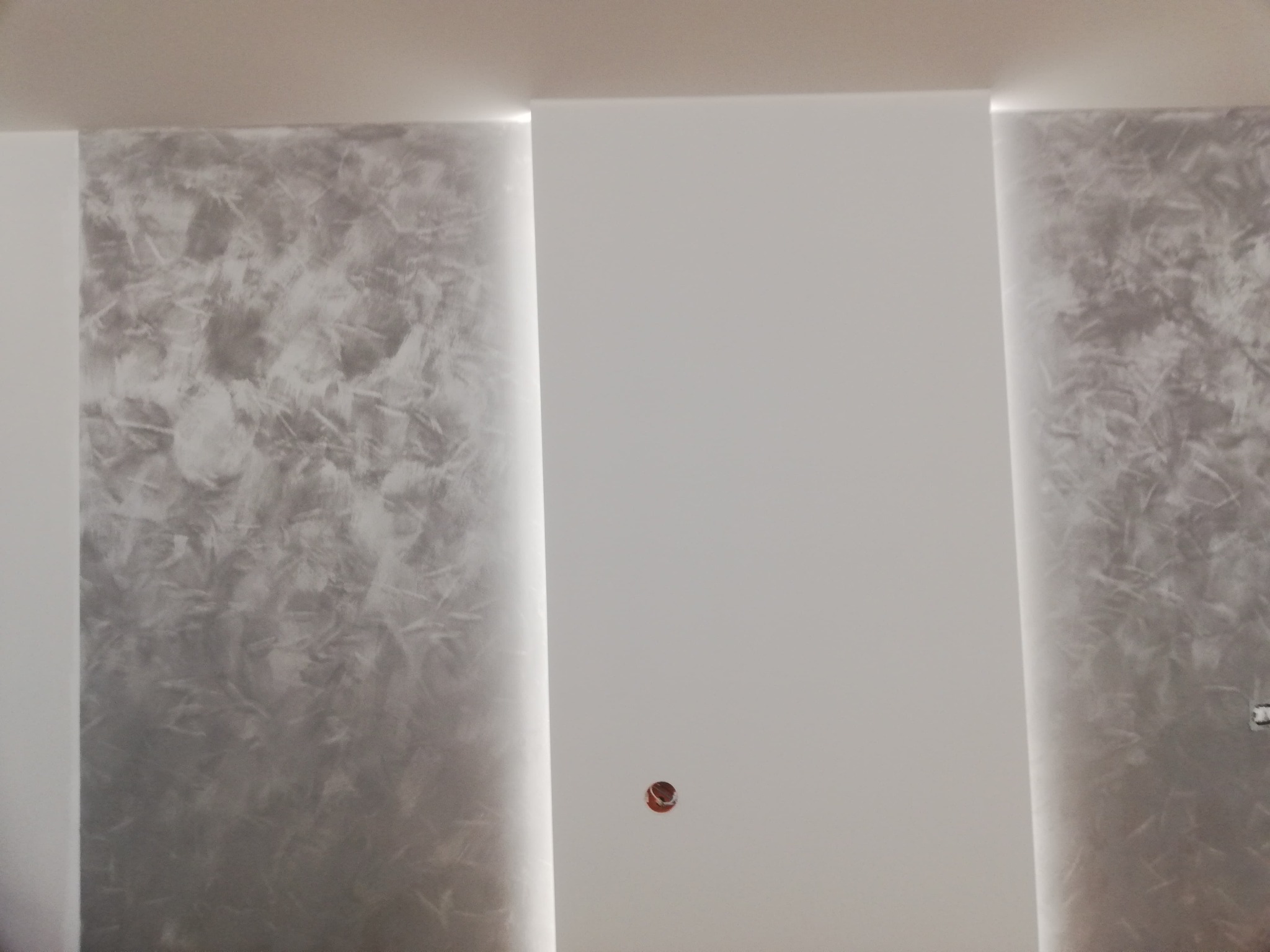 Fragment ściany z dekoracyjną, szarą strukturą tynku w pionowych pasach, oddzielonych gładką, białą powierzchnią. Widoczny otwór po gniazdku elektrycznym.