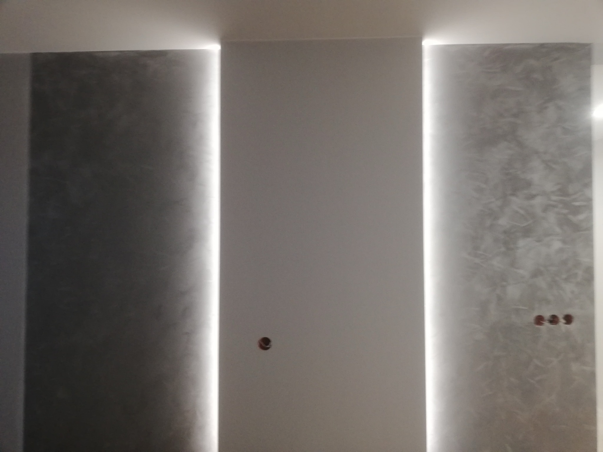 Ściana z dekoracyjnym tynkiem w odcieniach szarości, oświetlona pionowymi pasami LED, z widocznymi gniazdkami elektrycznymi. Minimalistyczny design wnętrza.