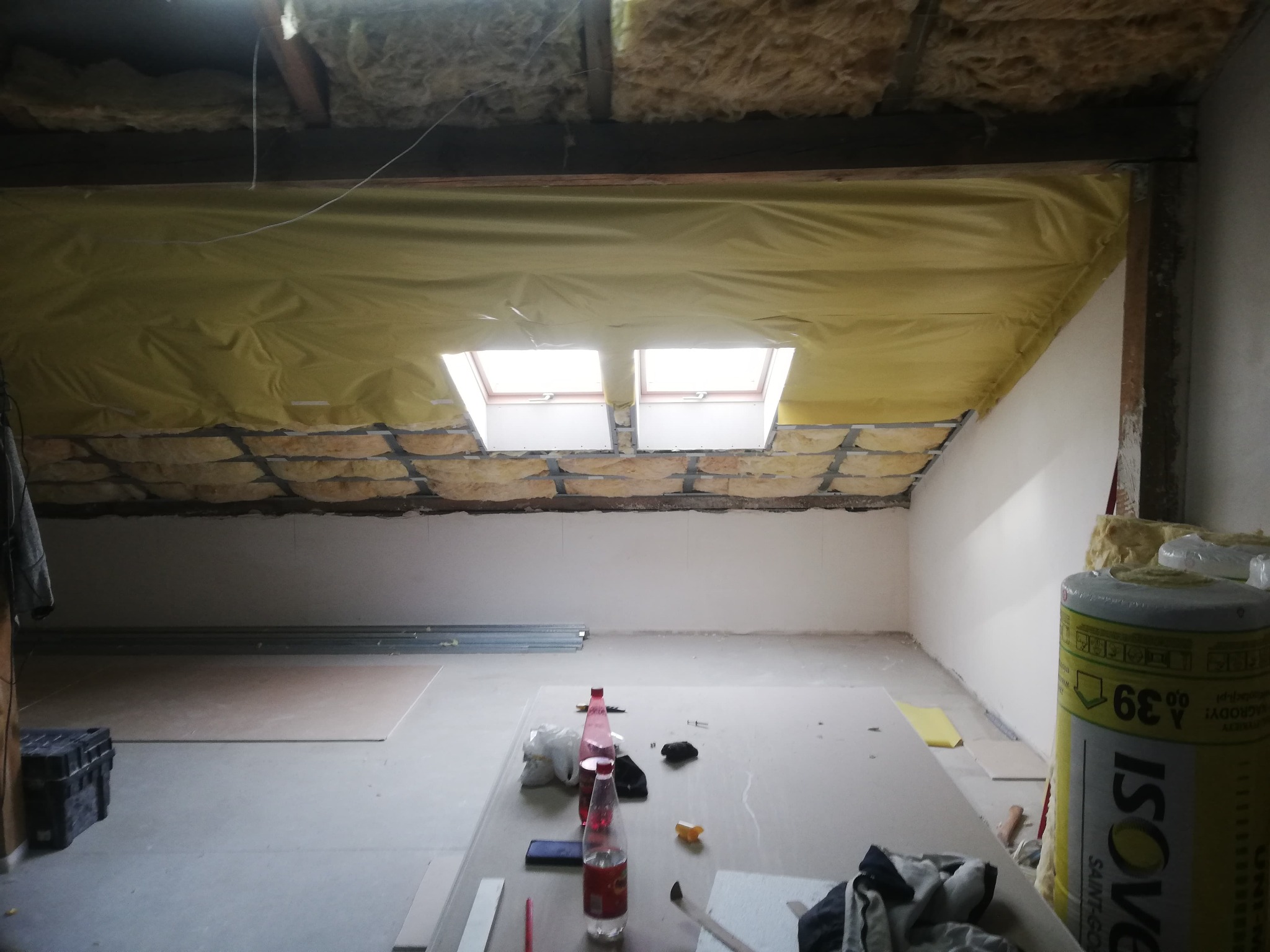 Poddasze w trakcie izolacji: widoczne okna dachowe, wełna mineralna, folia paroizolacyjna i materiały budowlane. Prace wykończeniowe w toku, surowe wnętrze.