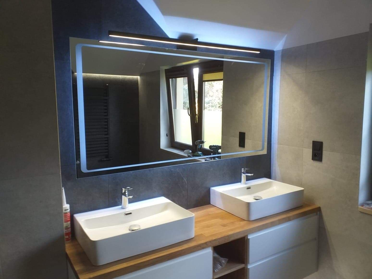 Nowoczesna łazienka z dwoma białymi umywalkami na drewnianym blacie, podświetlane lustro z widokiem na okno. Szare płytki na ścianach, minimalistyczny design.