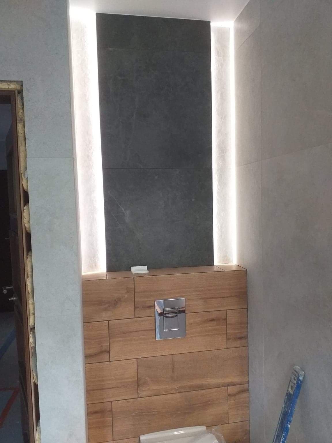 Nowoczesna łazienka: drewnopodobne płytki, szara ściana z podświetleniem LED i chromowany przycisk spłuczki. Widoczny fragment drabiny malarskiej.
