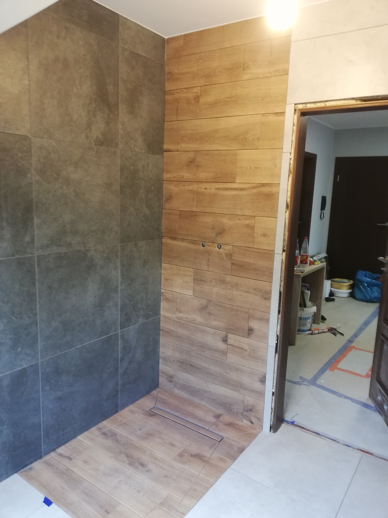 Wykończenie łazienki z szarymi płytkami i drewnopodobnymi panelami na ścianach. Widoczny odpływ liniowy w podłodze. Prace w toku, narzędzia w tle.