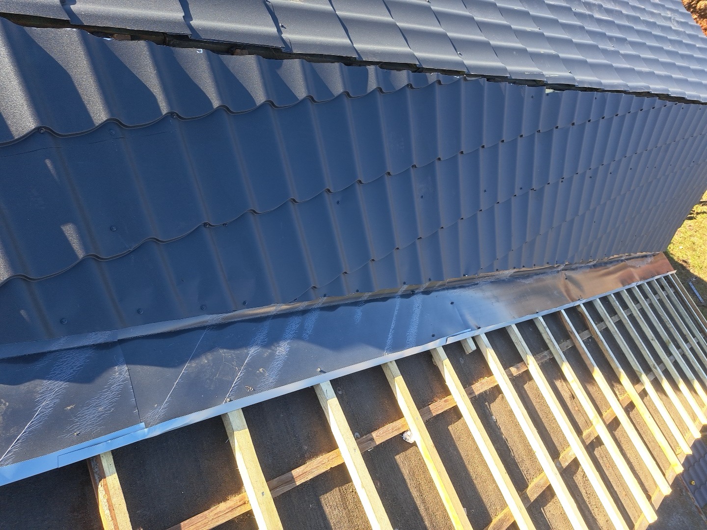 Fragment dachu z czarną dachówką karpiówką, widoczne elementy konstrukcji drewnianej i obróbki blacharskie. Ujęcie z góry, ostre światło.