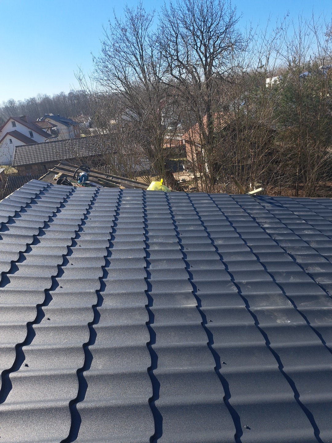 Nowy, grafitowy dach z blachodachówki modułowej, widok z góry na tle drzew i zabudowań w słoneczny dzień. Widoczne narzędzia dekarskie.
