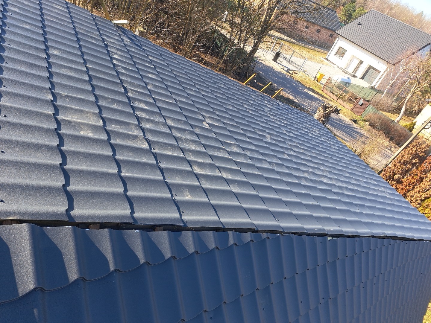 Szary dach z blachodachówki modułowej, widok z góry pod kątem. W tle budynki mieszkalne i drzewa bez liści. Widoczne mocowania blachy.