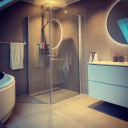 Gerard Wojewoda - Nowoczesna łazienka z kabiną prysznicową typu walk-in, okrągłym lustrem z podświetleniem LED i minimalistyczną szafką z umywalką w odcieniach szarości i błękitu.