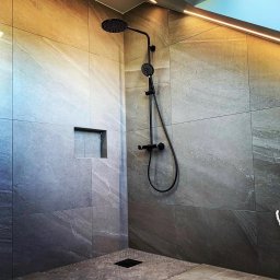 Gerard Wojewoda - Nowoczesna kabina prysznicowa z szarymi płytkami imitującymi kamień, czarną armaturą i wnęką w ścianie. Oświetlenie LED podkreśla fakturę płytek.