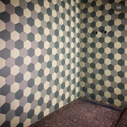 Gerard Wojewoda - Ściana i podłoga wyłożone heksagonalnymi płytkami w odcieniach szarości i beżu. Widoczne dwa otwory na rury w ścianie. Podłoga z ciemnego lastryko.