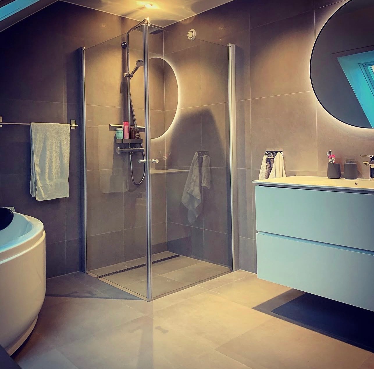 Nowoczesna łazienka z kabiną prysznicową typu walk-in, okrągłym lustrem z podświetleniem LED i minimalistyczną szafką z umywalką w odcieniach szarości i błękitu.