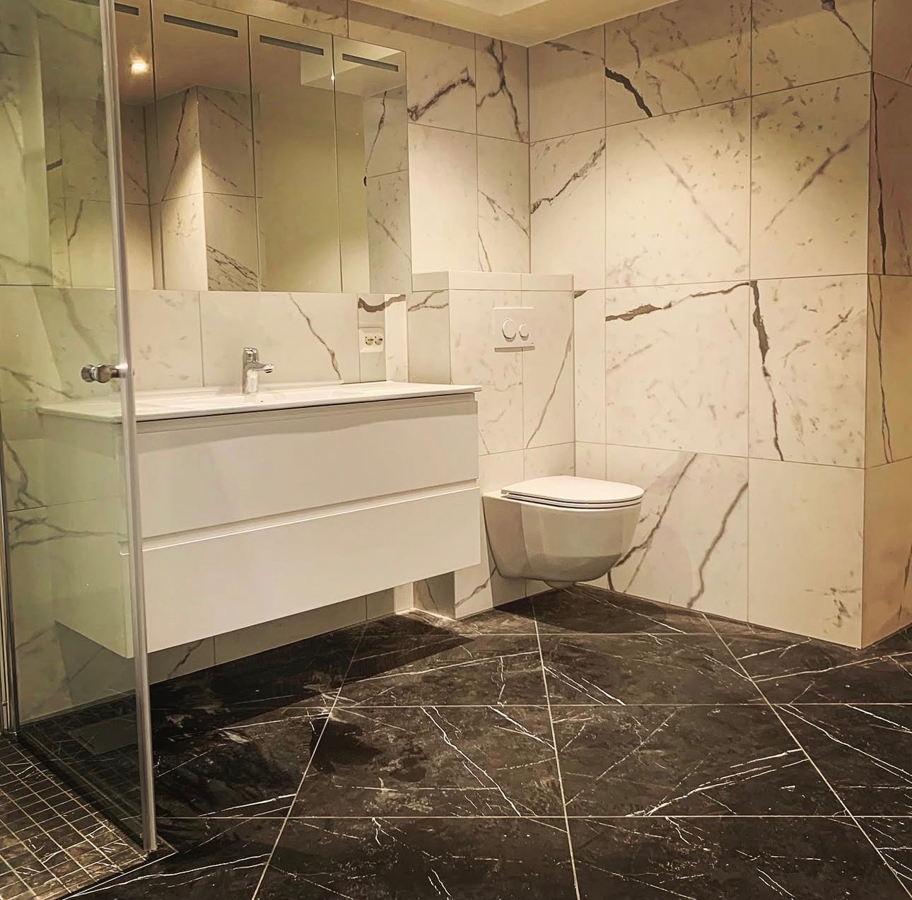 Nowoczesna łazienka z marmurowymi płytkami na ścianach i podłodze. Biała szafka pod umywalką i wisząca toaleta. Kabina prysznicowa ze szklanymi drzwiami.