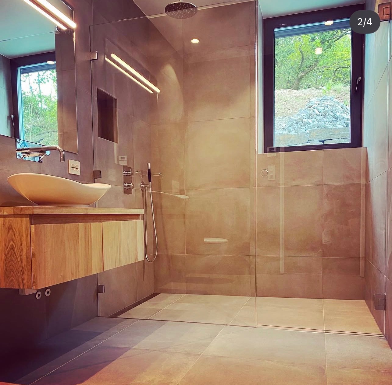 Nowoczesna łazienka z kabiną prysznicową typu walk-in, drewnianą szafką pod umywalką i oknem z widokiem na zieleń. Minimalistyczny design, szare płytki i naturalne światło.