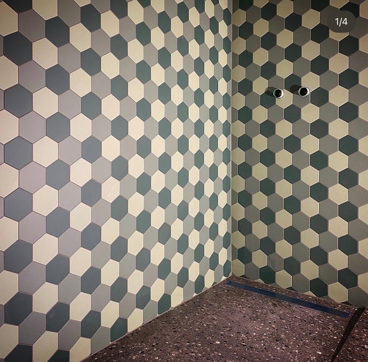 Ściana i podłoga wyłożone heksagonalnymi płytkami w odcieniach szarości i beżu. Widoczne dwa otwory na rury w ścianie. Podłoga z ciemnego lastryko.