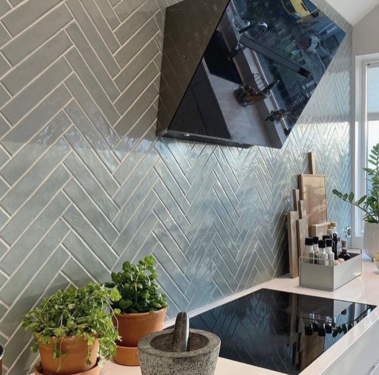 Ściana w kuchni wyłożona błyszczącymi, szarymi płytkami ułożonymi w jodełkę, z czarnym okapem i roślinami w doniczkach na blacie. Widoczne przyprawy i deski do krojenia.