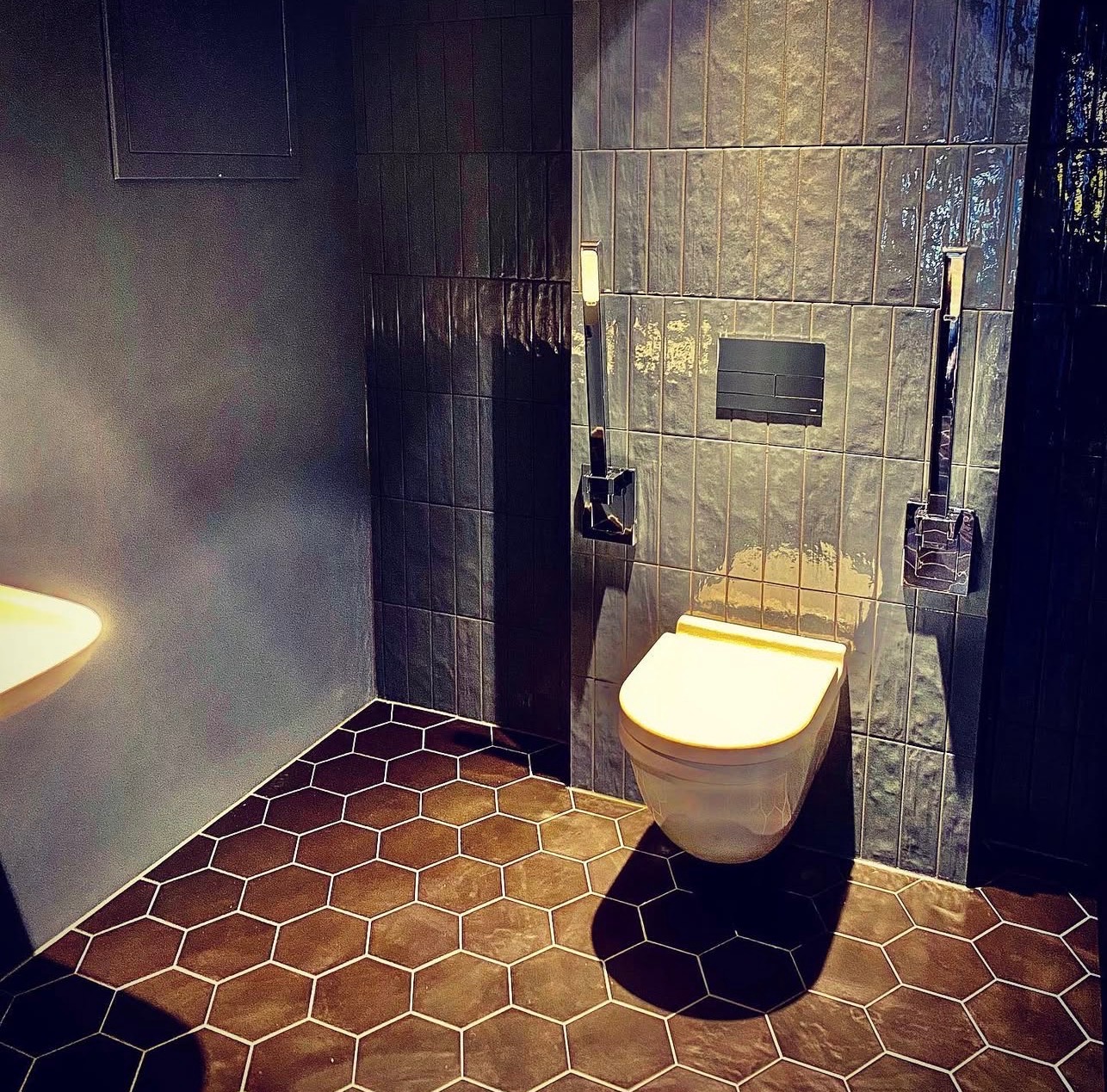 Nowoczesna łazienka z heksagonalną podłogą i ciemnymi płytkami ściennymi. Wisząca toaleta z chromowanymi akcesoriami. Minimalistyczny design i stonowana kolorystyka.