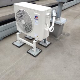 Kamil Zimorski - Total Klima - Biała jednostka zewnętrzna klimatyzacji GREE zamontowana na dachu budynku, widoczne elementy mocujące i rury. Szare tło dachu i fragment budynku w tle.