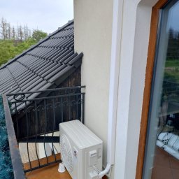 Kamil Zimorski - Total Klima - Kompaktowa jednostka zewnętrzna klimatyzacji GREE na balkonie z brązowymi płytkami, obok okna z drewnianą ramą i białej rury maskującej. Widok na dach i zieleń.