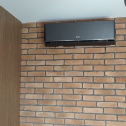 Kamil Zimorski - Total Klima - Grafitowy klimatyzator marki Gree zamontowany na ścianie z cegły dekoracyjnej. Widoczny fragment drewnianej zabudowy. Minimalistyczny design.