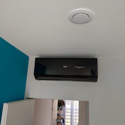 Kamil Zimorski - Total Klima - Czarny klimatyzator Haier na białej ścianie z turkusowym akcentem, widoczny okrągły nawiewnik sufitowy i odbicie wnętrza pokoju w lustrzanej szafie.