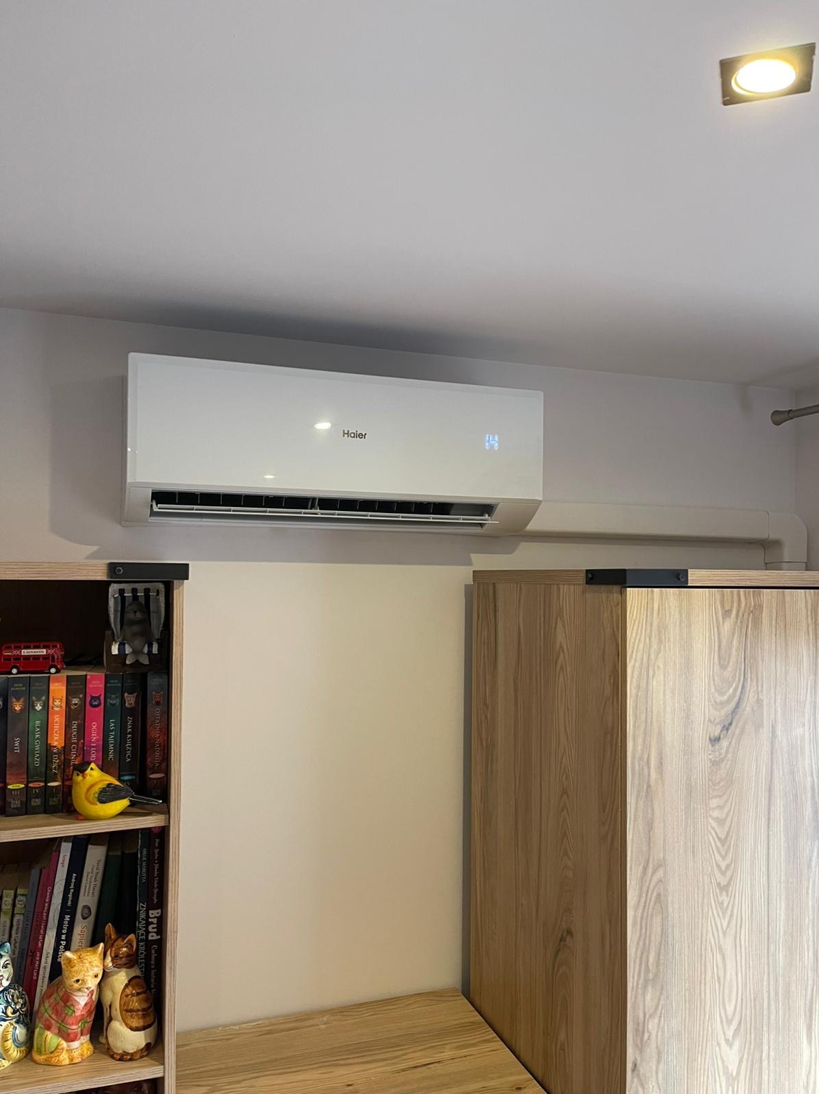 Biały klimatyzator Haier zamontowany na ścianie w pokoju, widoczny fragment regału z książkami i dekoracjami. Minimalistyczny design, oświetlenie punktowe.