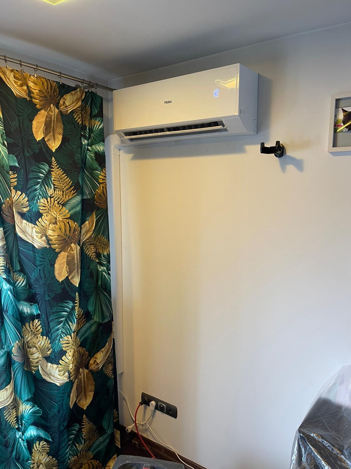 Biały klimatyzator Haier zamontowany na ścianie w pokoju z zasłoną w tropikalne wzory. Widoczne gniazdka elektryczne i wieszak na ścianie.