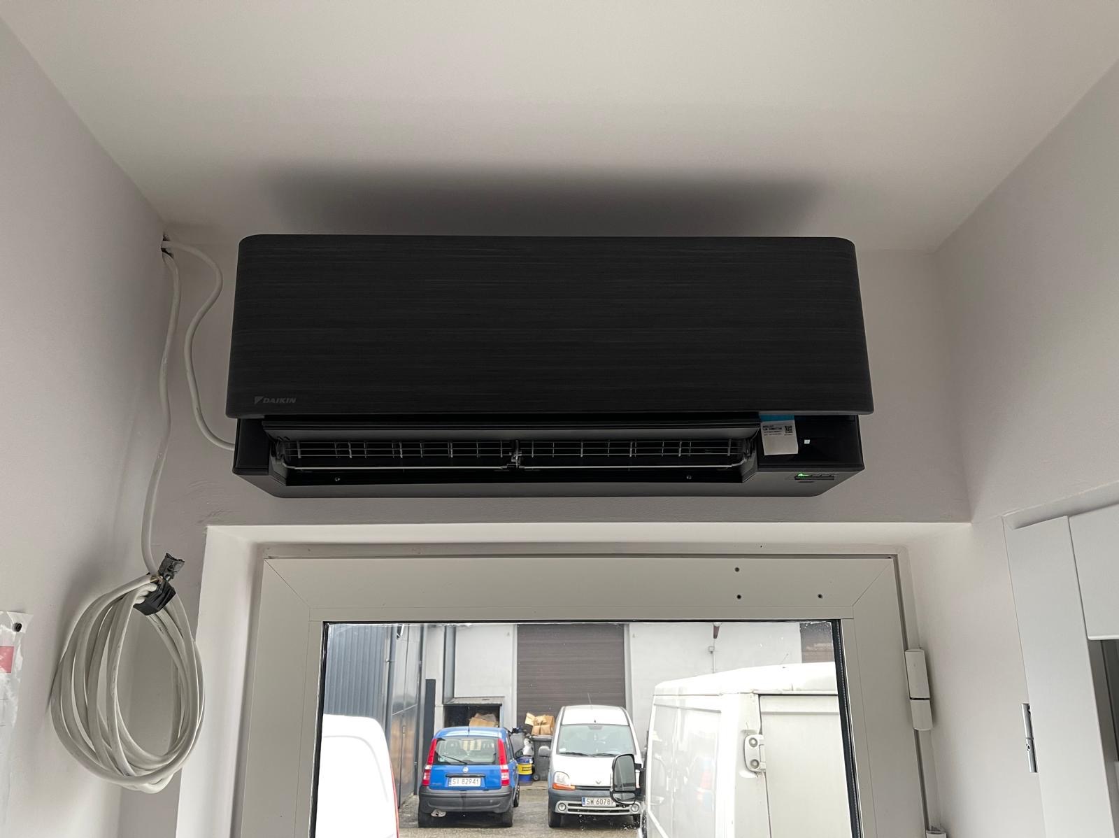 Czarny klimatyzator Daikin zamontowany pod sufitem, widoczne kable i okno z widokiem na zaparkowane samochody. Minimalistyczny design i nowoczesne wykończenie.