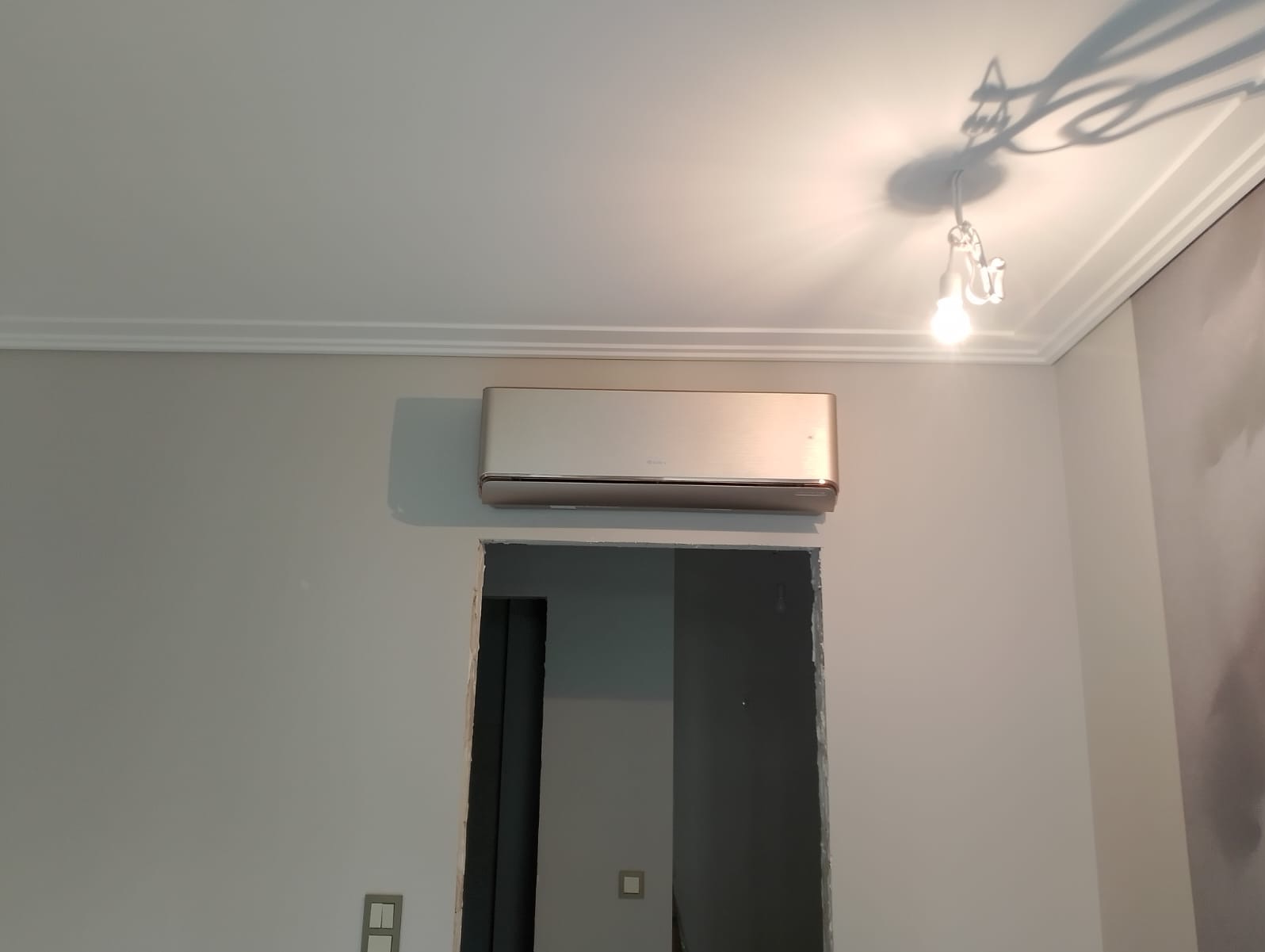 Złoty klimatyzator na szarej ścianie nad otworem drzwiowym. Widoczne listwy sufitowe i lampa. Ujęcie z dołu, podkreślające wysokość pomieszczenia.