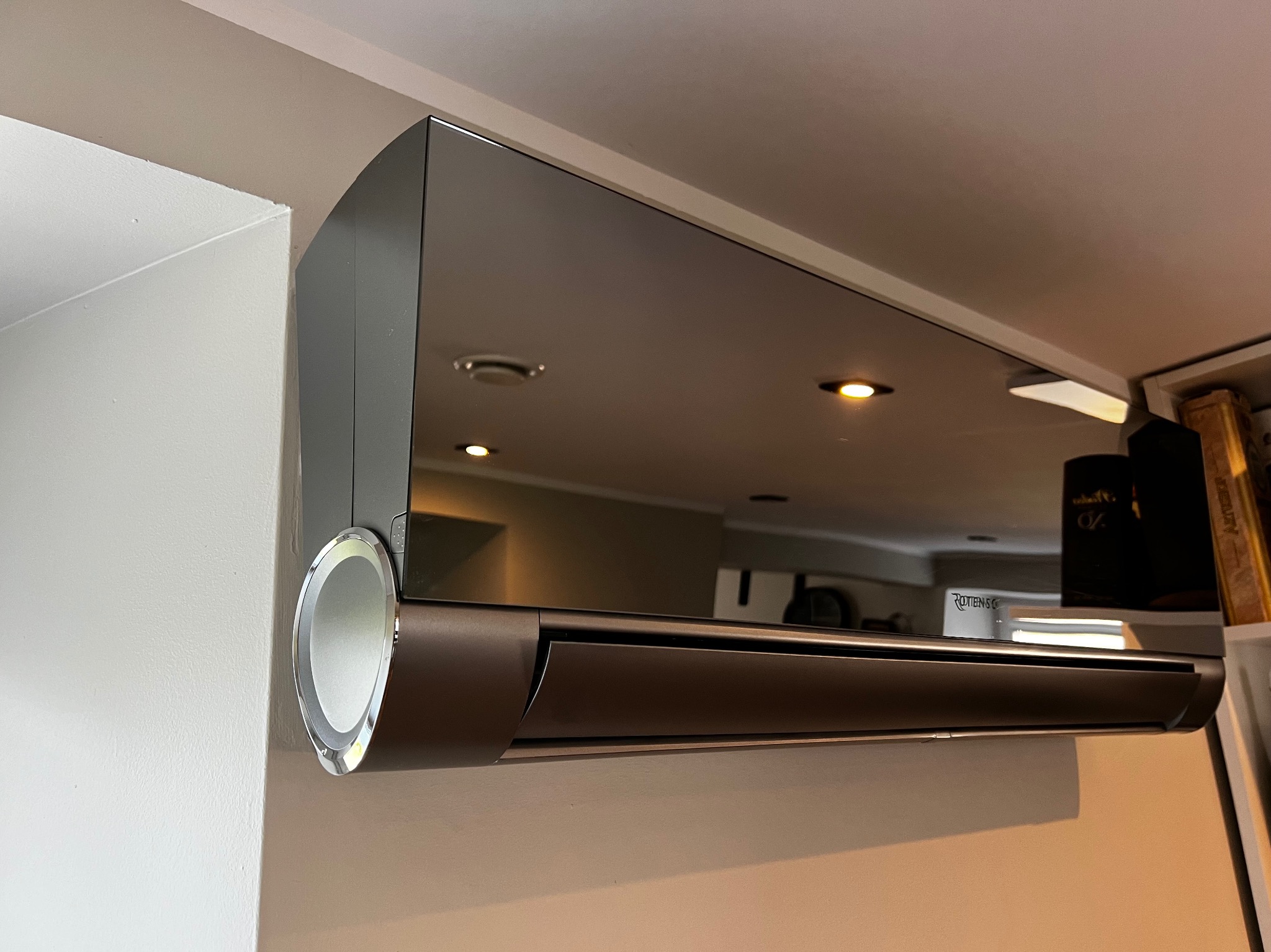 Nowoczesny klimatyzator ścienny w odcieniach szarości i brązu, zamontowany w pomieszczeniu. Widoczne odbicie wnętrza w lustrzanej obudowie urządzenia.