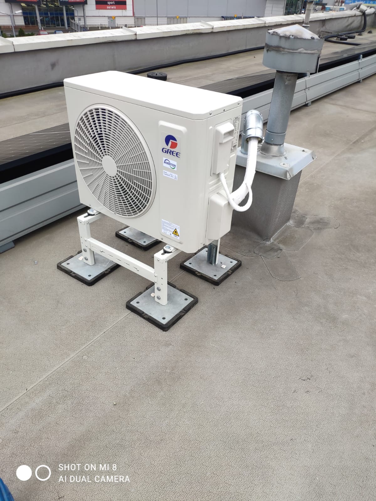 Biała jednostka zewnętrzna klimatyzacji GREE zamontowana na dachu budynku, widoczne elementy mocujące i rury. Szare tło dachu i fragment budynku w tle.