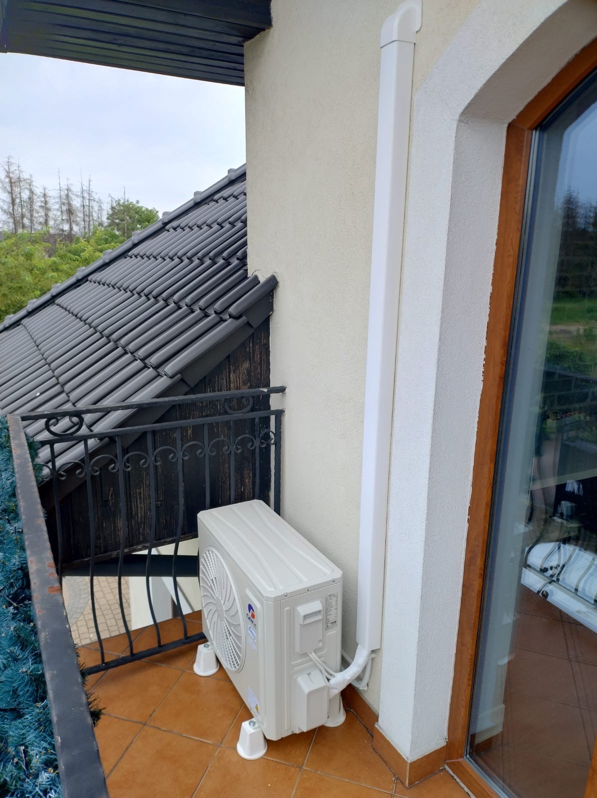 Kompaktowa jednostka zewnętrzna klimatyzacji GREE na balkonie z brązowymi płytkami, obok okna z drewnianą ramą i białej rury maskującej. Widok na dach i zieleń.
