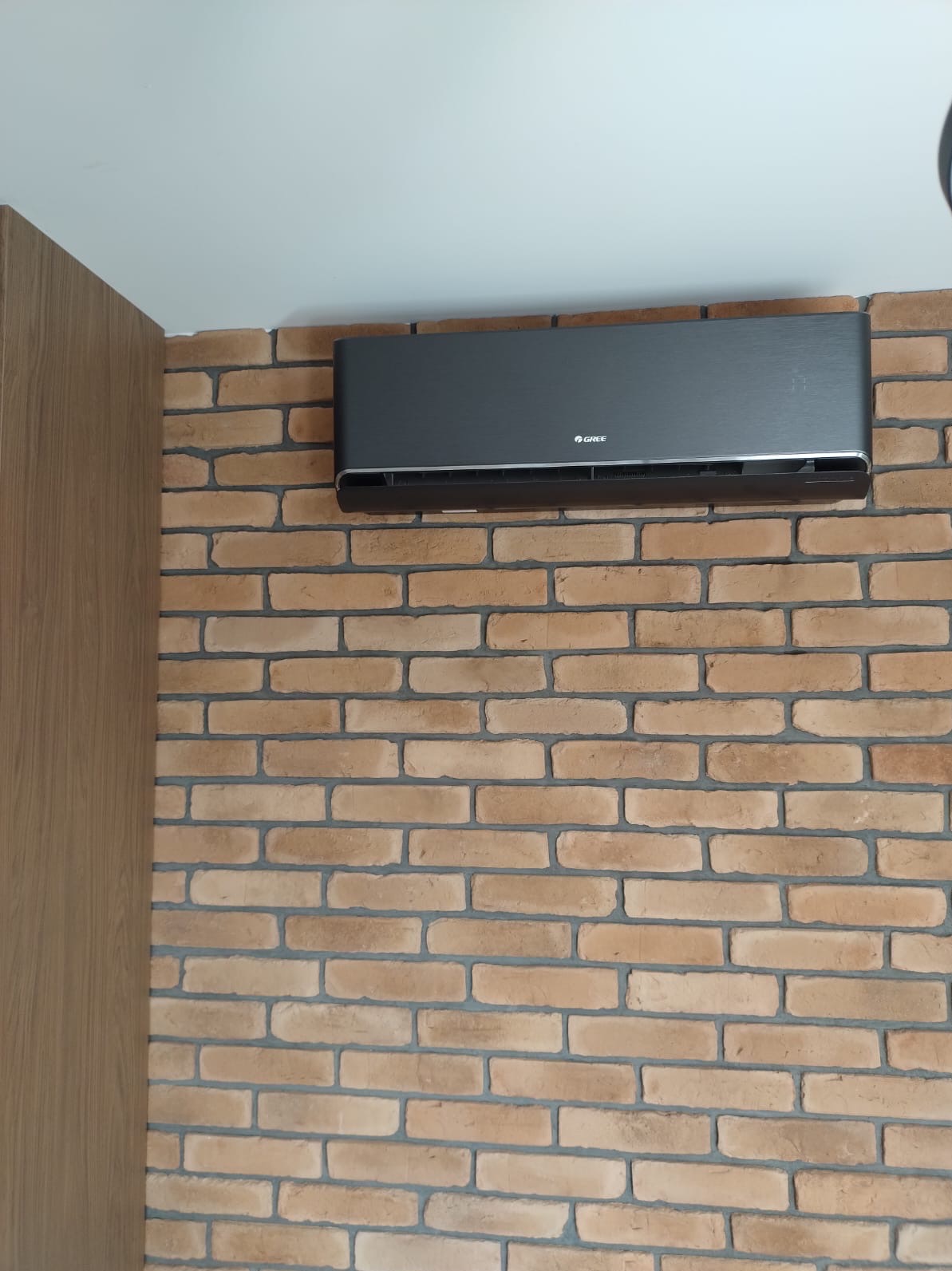 Grafitowy klimatyzator marki Gree zamontowany na ścianie z cegły dekoracyjnej. Widoczny fragment drewnianej zabudowy. Minimalistyczny design.