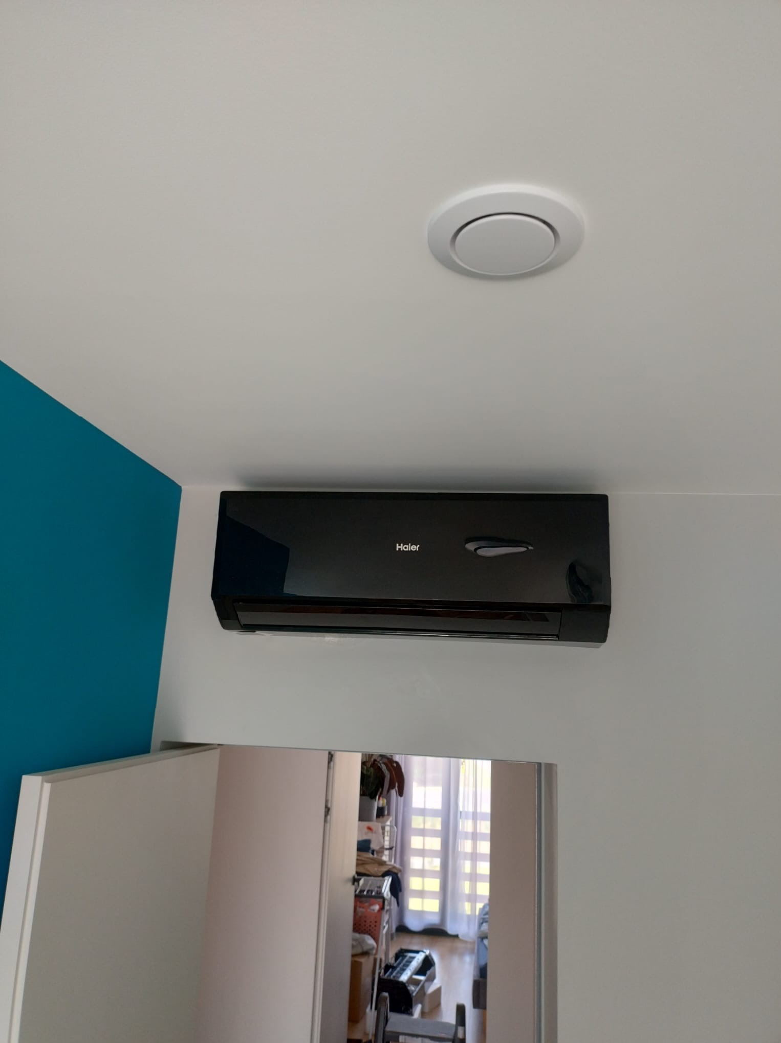 Czarny klimatyzator Haier na białej ścianie z turkusowym akcentem, widoczny okrągły nawiewnik sufitowy i odbicie wnętrza pokoju w lustrzanej szafie.