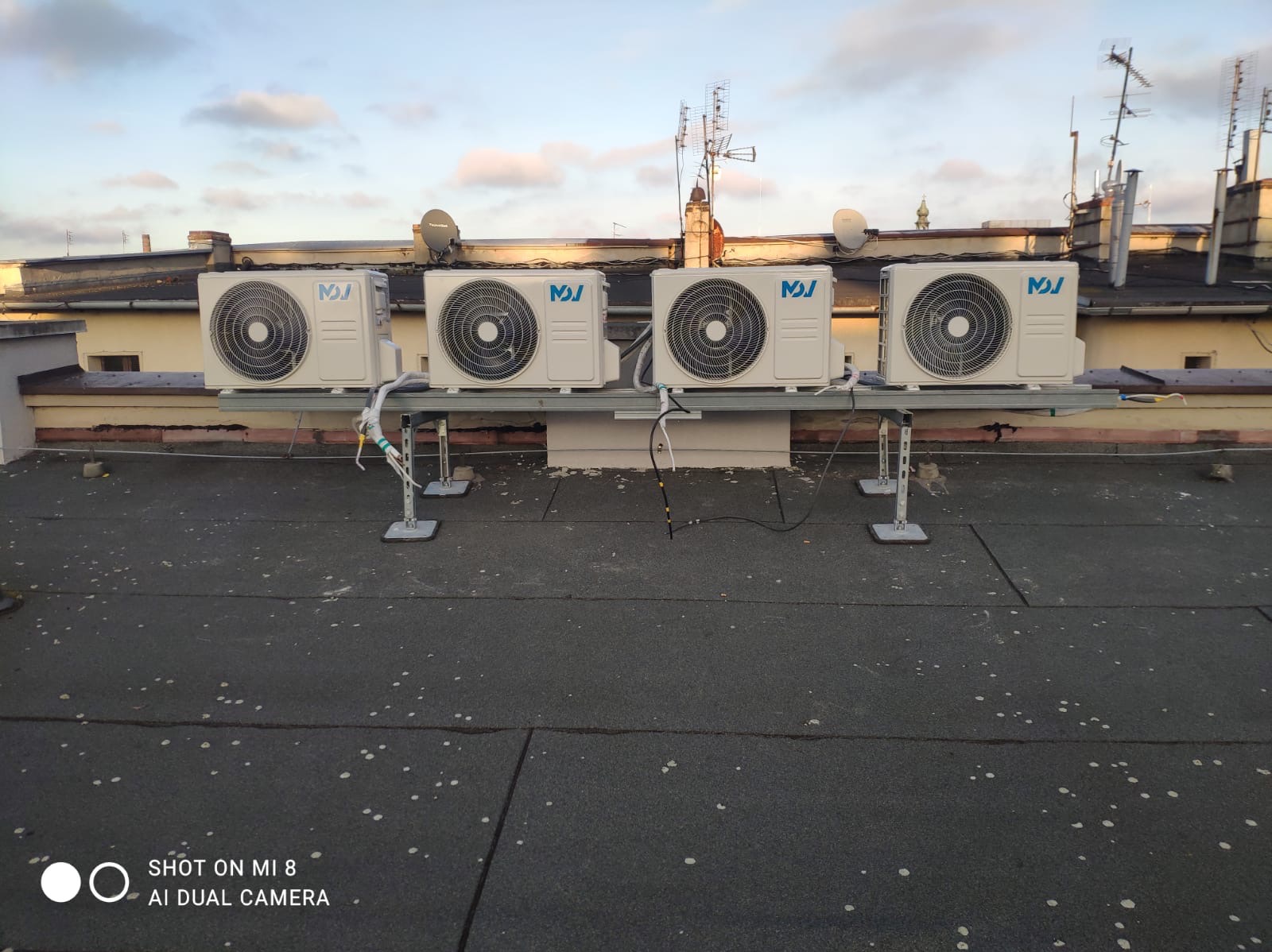 Cztery klimatyzatory na dachu budynku, zamontowane na metalowej konstrukcji. W tle widoczne inne budynki i anteny.