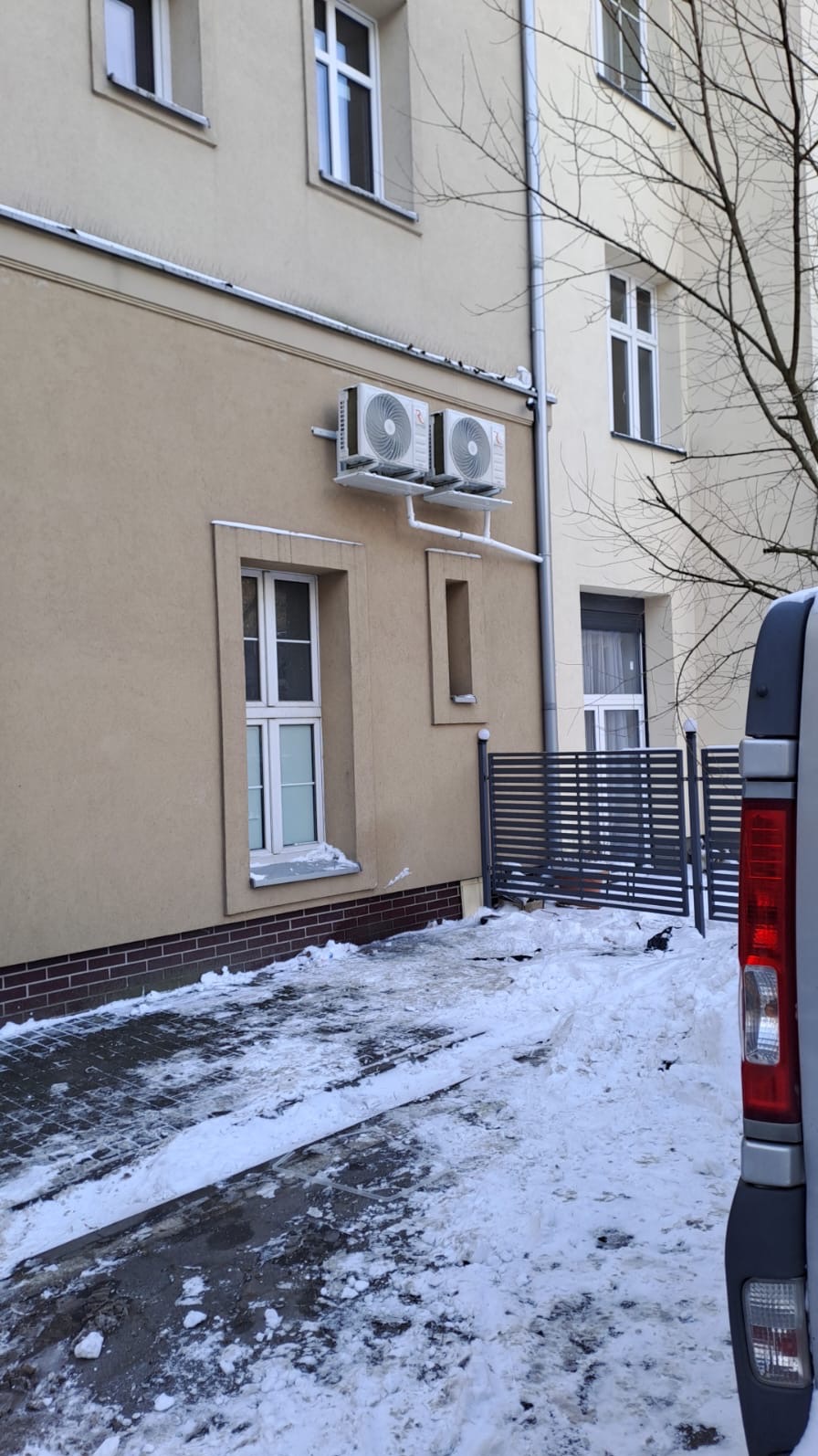 Dwa białe klimatyzatory zamontowane na elewacji budynku w zimowej scenerii. Widoczne rury odprowadzające skropliny. Część samochodu dostawczego w kadrze.