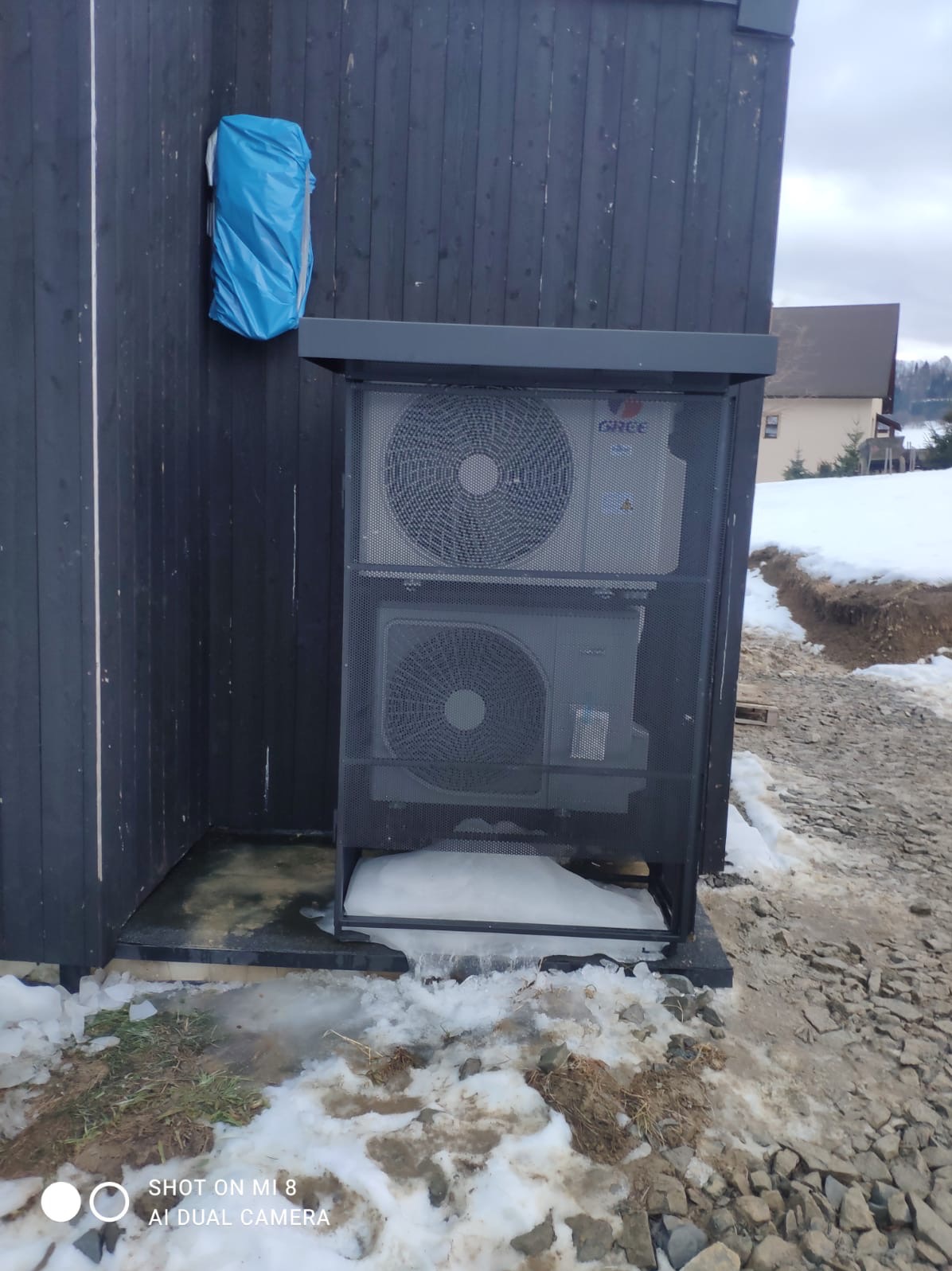 Zewnętrzna jednostka klimatyzacji GREE, zabezpieczona metalową osłoną, zamontowana przy ciemnej, drewnianej elewacji budynku w zimowej scenerii. Widoczny niebieski pokrowiec.