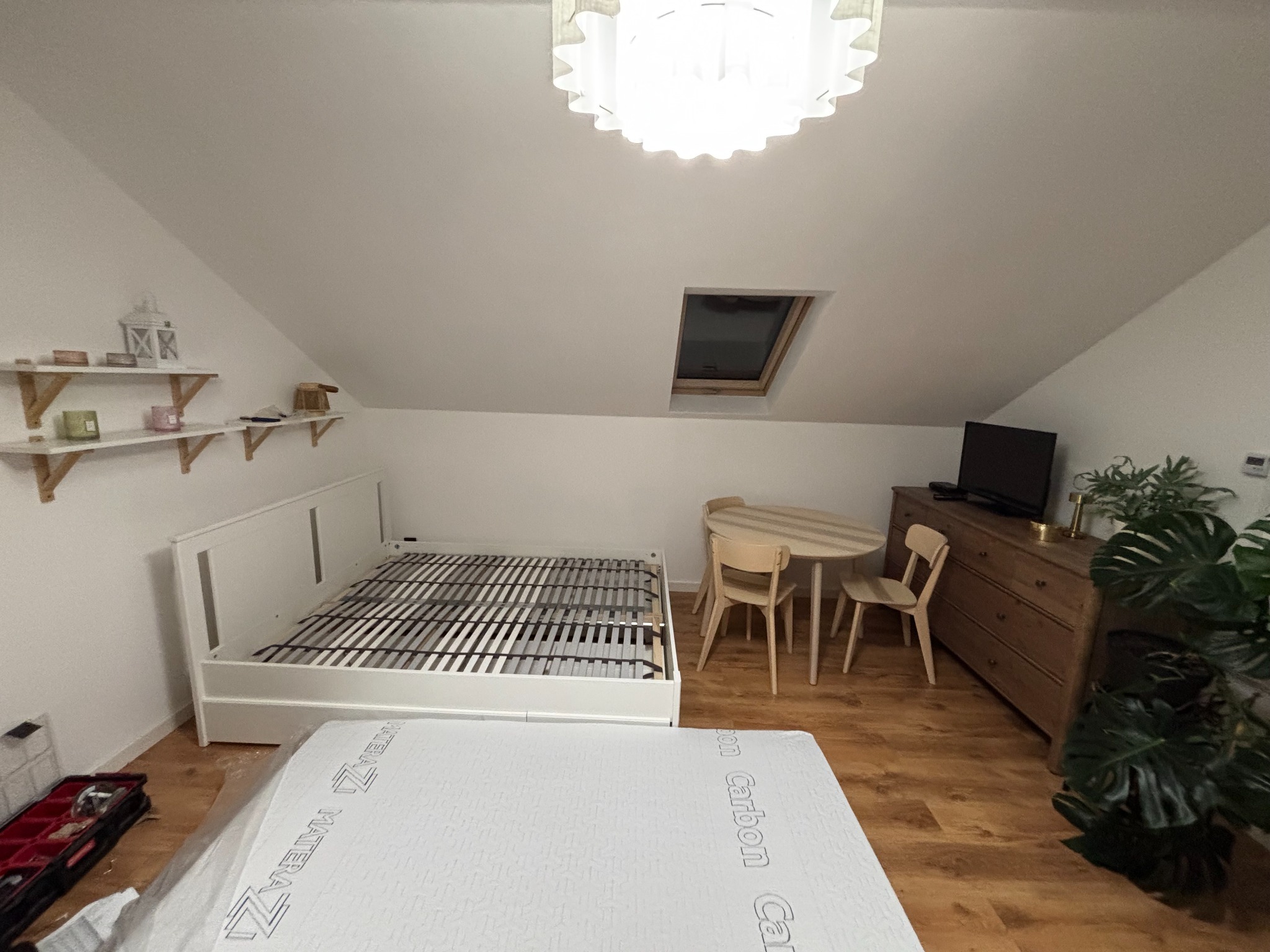 Pokój na poddaszu z częściowo złożonym białym łóżkiem, stolikiem i komodą. Na podłodze leży materac w folii. Widoczne narzędzia i półki z dekoracjami.