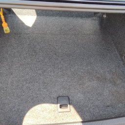 KingClean Expert - Wnętrze bagażnika samochodu wyłożone szarą wykładziną dywanową. Widoczne elementy mocujące i uchwyt. Jasne plamy światła podkreślają strukturę materiału.